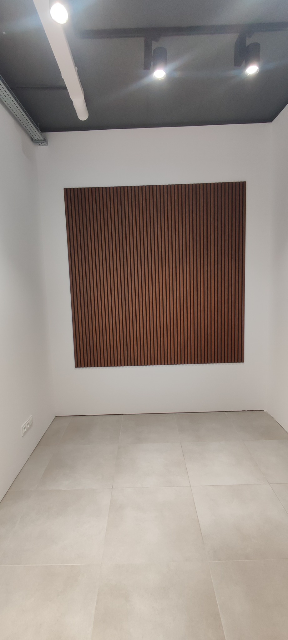 Pusty pokój z beżową podłogą wyłożoną płytkami i dekoracyjnym panelem ściennym z pionowych, brązowych listewek na białej ścianie; sufit z czarnymi reflektorami.