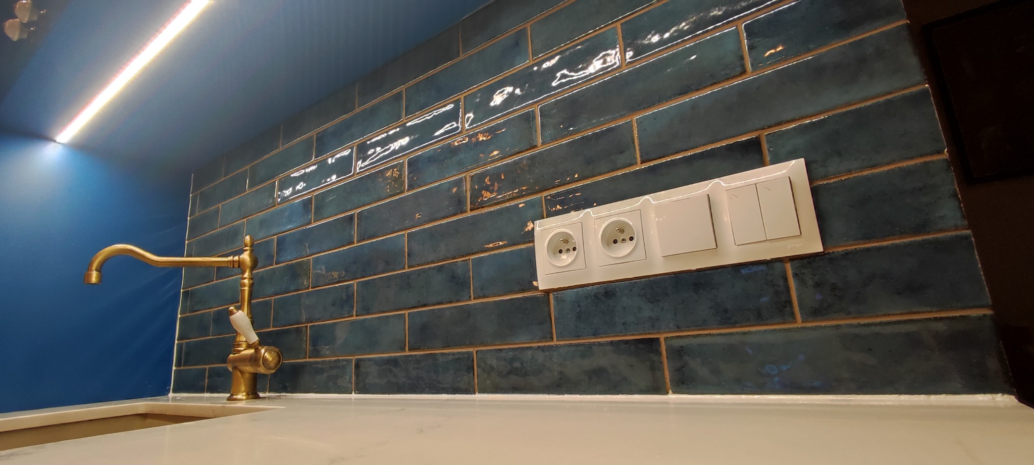 Błyszczące, ciemnoniebieskie płytki ułożone w cegiełkę na ścianie nad blatem kuchennym, z baterią w stylu retro z elementami porcelany, oraz gniazdkami i włącznikami światła w białej ramce.