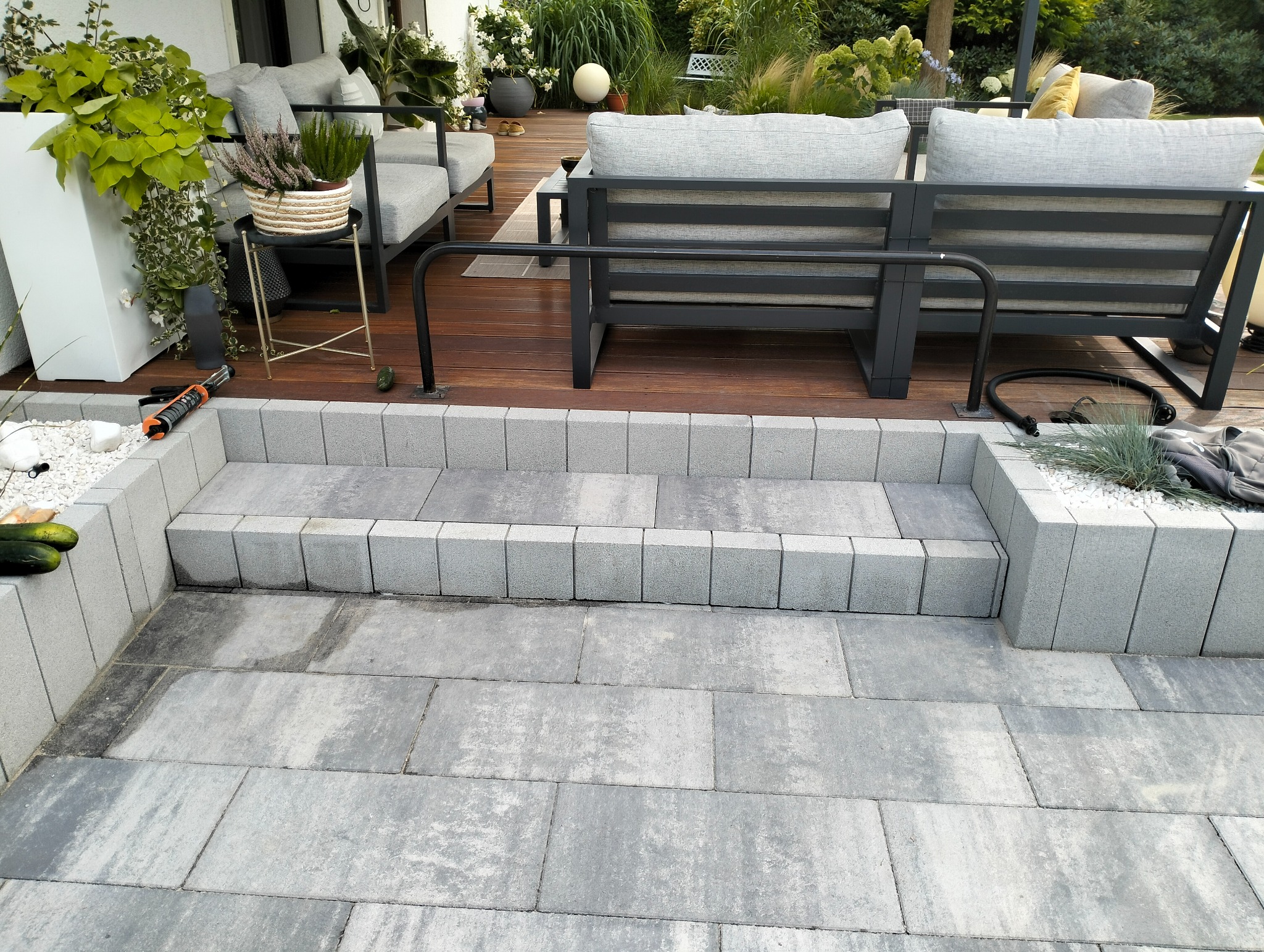 Wykończony taras z kostki brukowej w odcieniach szarości, z niskimi schodami i elementami małej architektury, takimi jak donice i meble ogrodowe. Na schodach widoczne narzędzie do fugowania. W tle...