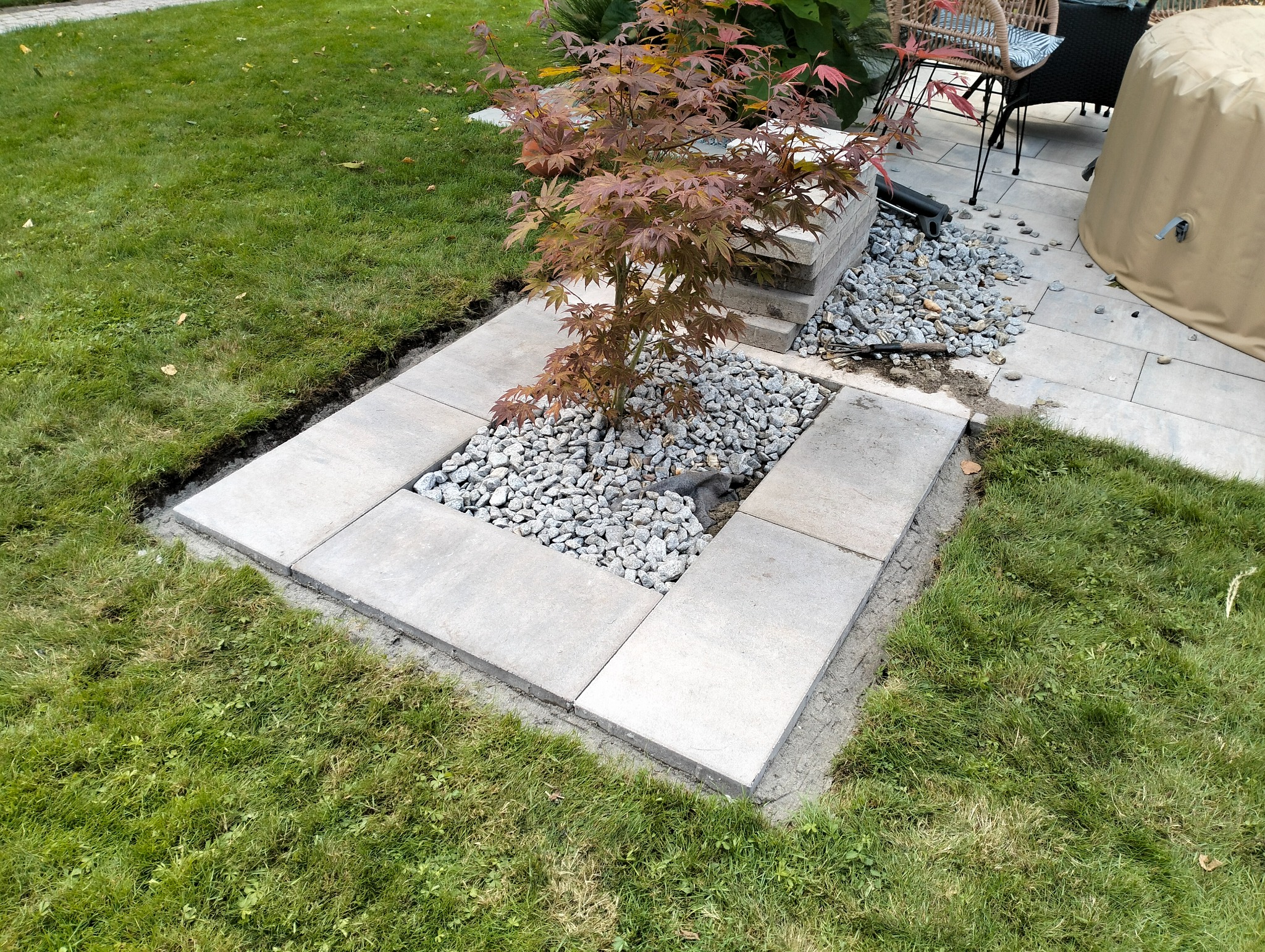 Obrzeże z jasnoszarych płyt wokół rabaty z ozdobnym drzewkiem i białymi kamykami, widoczne ślady świeżo wykonanych prac ziemnych na trawniku.