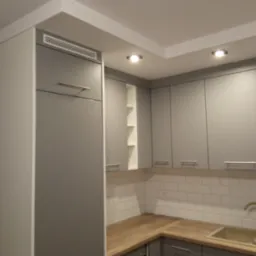 Szare szafki kuchenne z drewnianym blatem i złotą baterią, białe płytki na ścianie, oświetlenie punktowe w suficie.