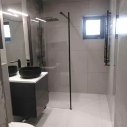 Nowoczesna łazienka z kabiną prysznicową typu walk-in, czarną armaturą i umywalką nablatową, wykończona płytkami w odcieniach szarości, z widocznym fragmentem toalety.