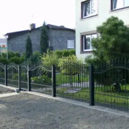 Czarny metalowy płot z ozdobnymi elementami, oddzielający posesję z domem i zielenią od drogi gruntowej, z widocznym w tle banerem reklamowym.