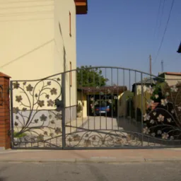 Otwarta, metalowa brama wjazdowa z dekoracyjnym motywem liści i winorośli, zamontowana na ceglanych słupkach, z widokiem na podjazd i zaparkowany samochód.