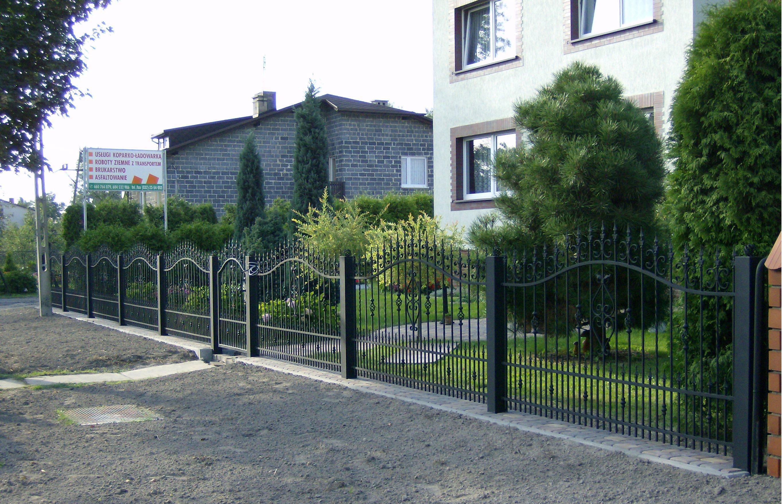 Czarny metalowy płot z ozdobnymi elementami, oddzielający posesję z domem i zielenią od drogi gruntowej, z widocznym w tle banerem reklamowym.