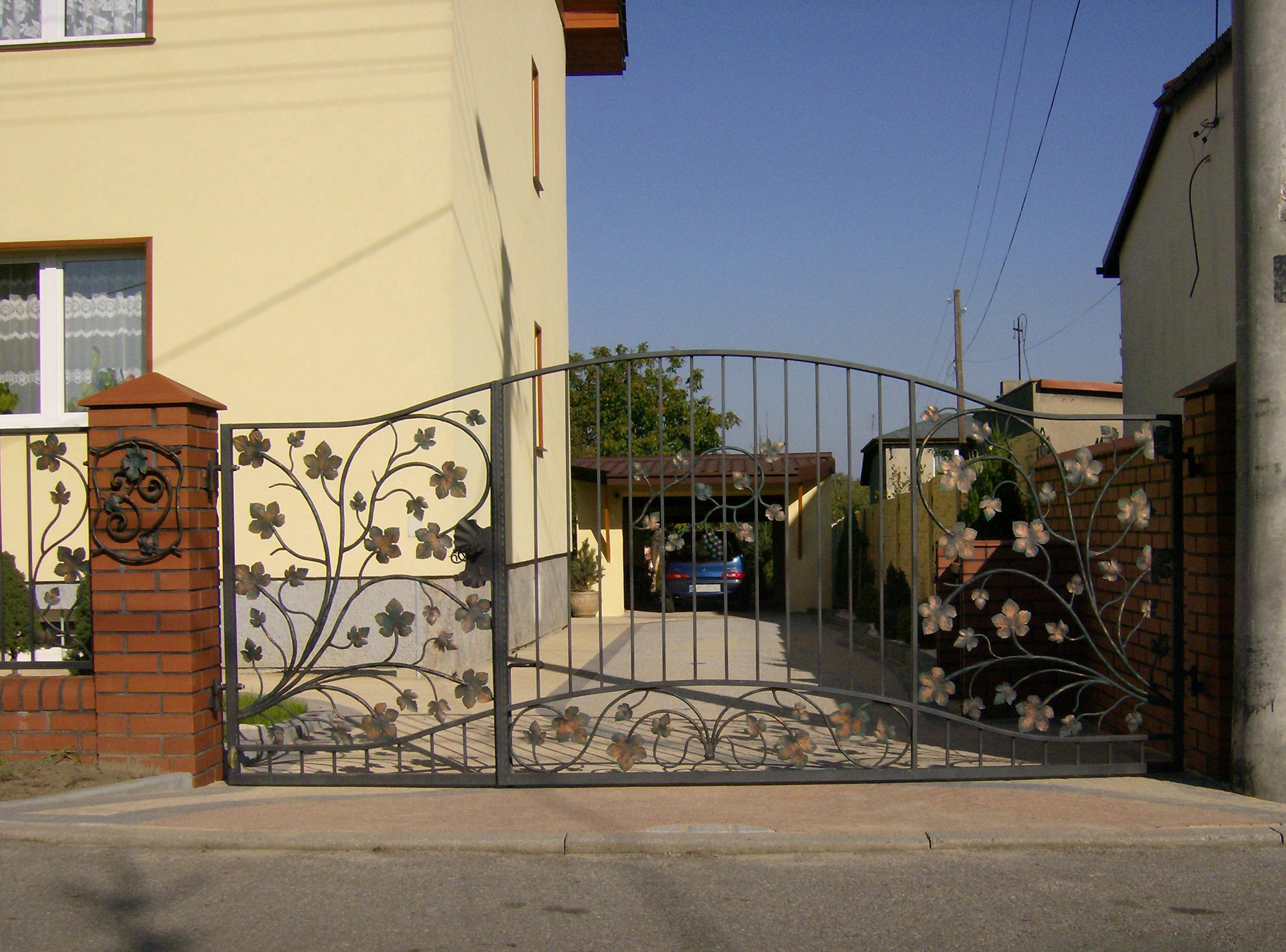 Otwarta, metalowa brama wjazdowa z dekoracyjnym motywem liści i winorośli, zamontowana na ceglanych słupkach, z widokiem na podjazd i zaparkowany samochód.