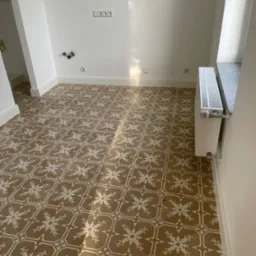 Wnętrze po remoncie z wzorzystą, brązowo-białą podłogą z płytek ceramicznych, białe ściany z widocznymi gniazdkami i grzejnikiem, światło słoneczne padające na podłogę.