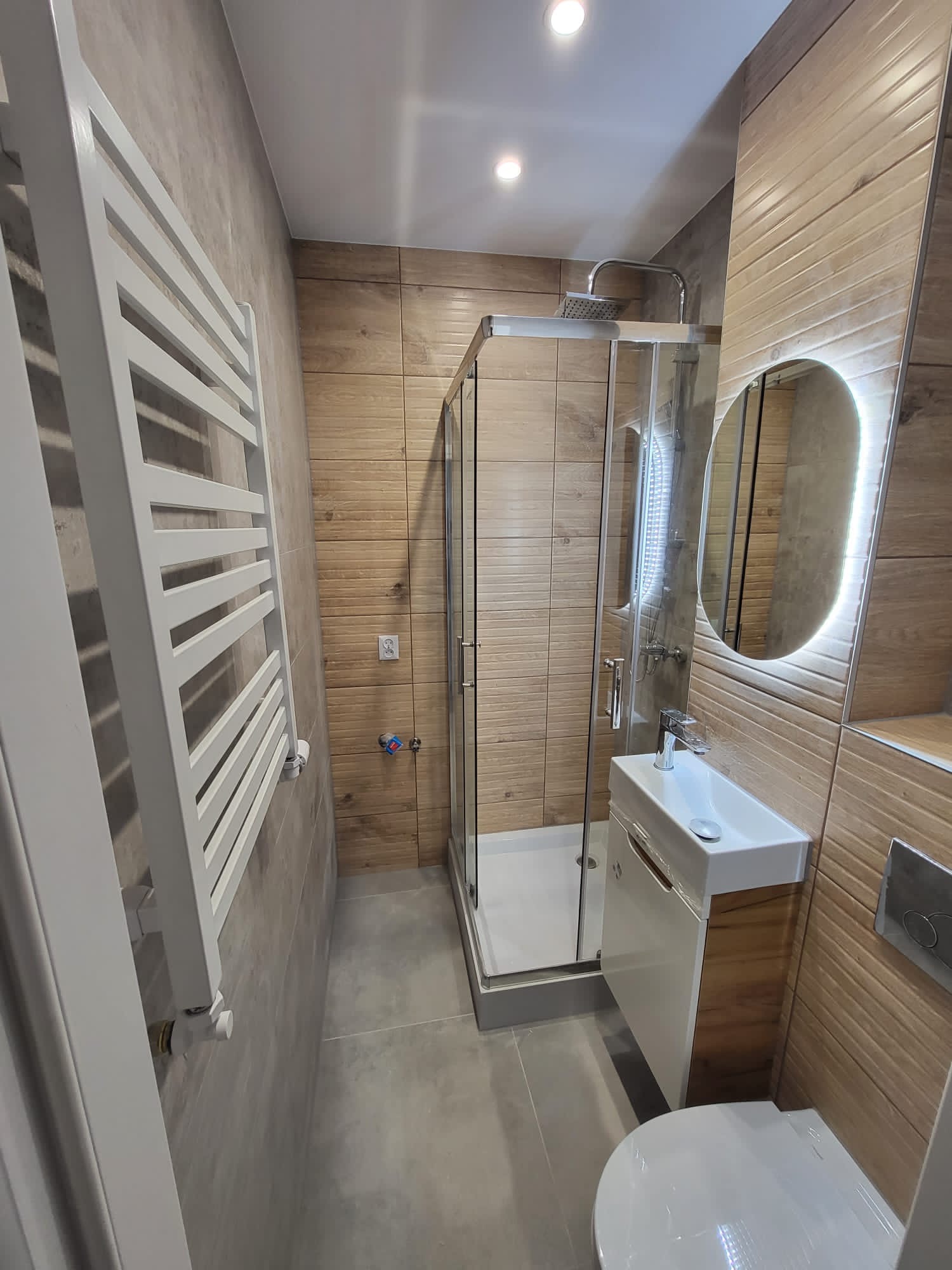 Nowoczesna łazienka z kabiną prysznicową, umywalką nablatową na szafce z drewnopodobnym frontem, podświetlanym lustrem owalnym i białym grzejnikiem drabinkowym na ścianie wyłożonej płytkami...