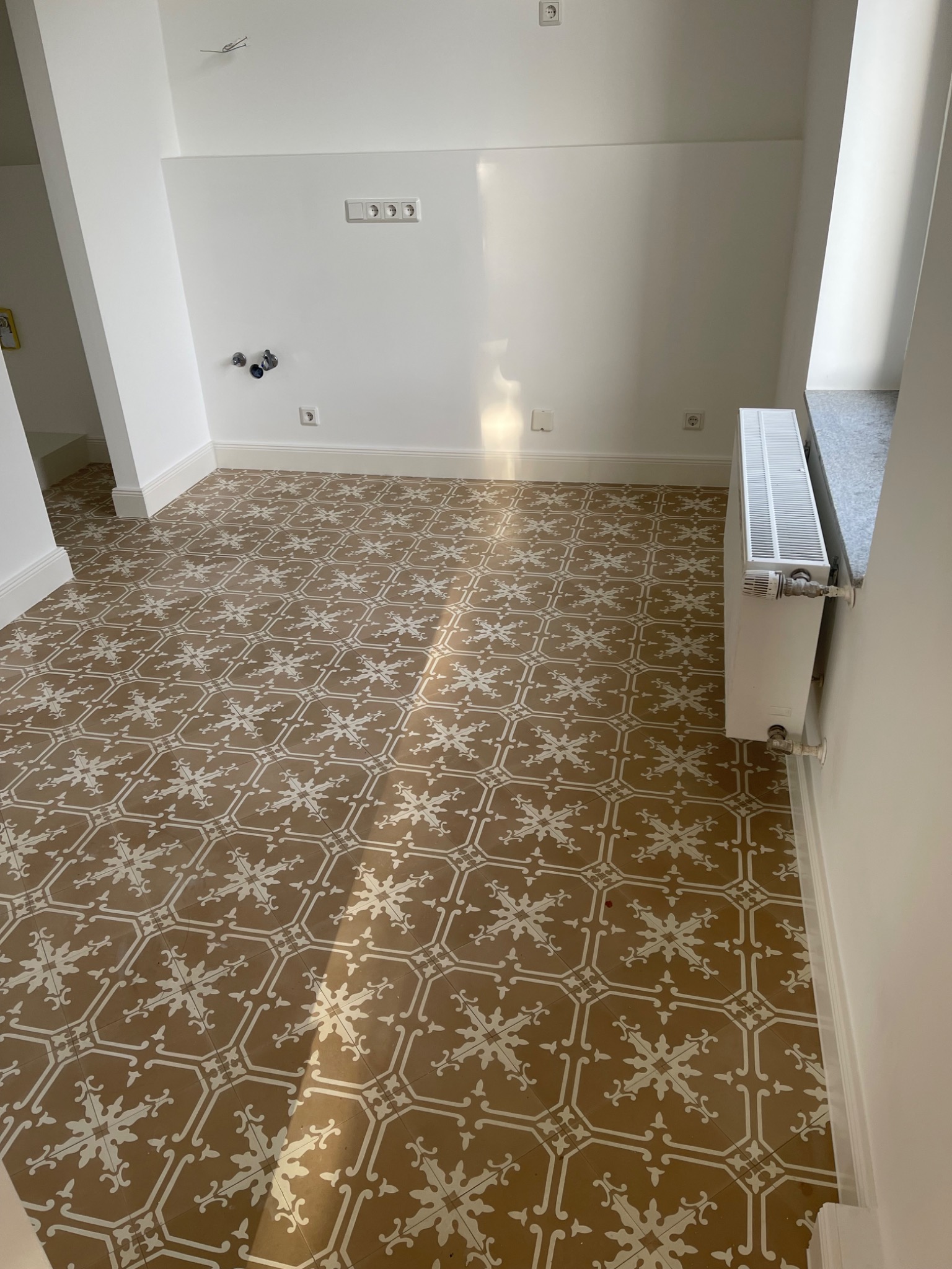 Wnętrze po remoncie z wzorzystą, brązowo-białą podłogą z płytek ceramicznych, białe ściany z widocznymi gniazdkami i grzejnikiem, światło słoneczne padające na podłogę.