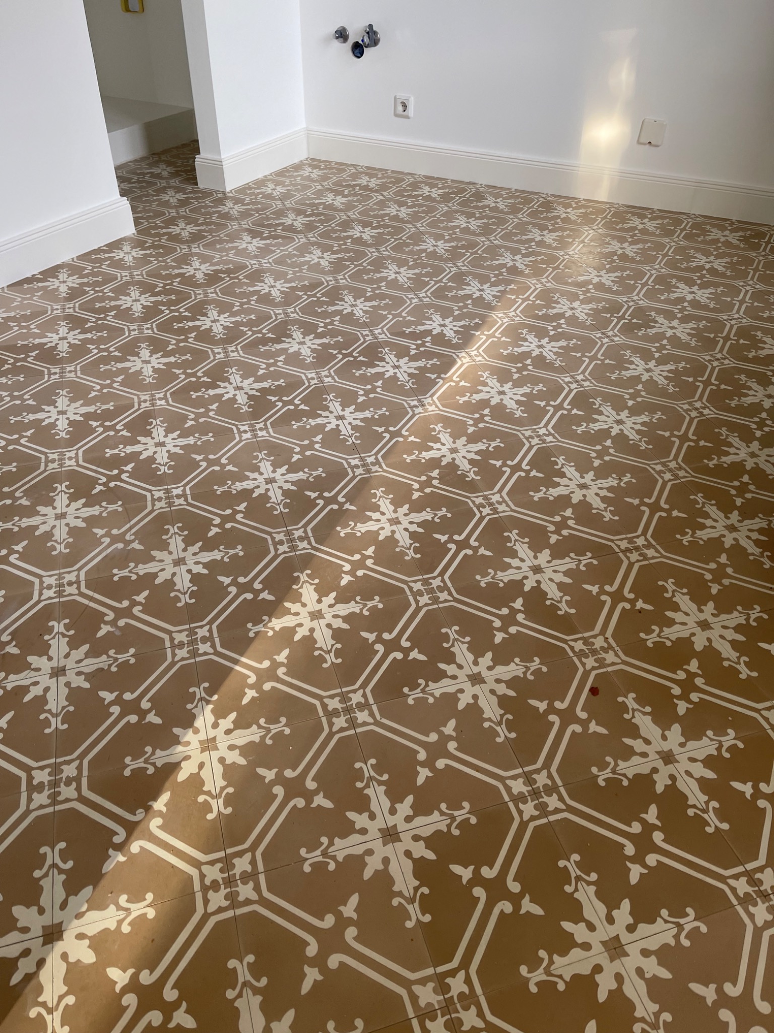 Podłoga wyłożona brązowymi płytkami z białym wzorem w kształcie kwiatów i geometrycznych elementów, widoczne gniazdka elektryczne i listwy przypodłogowe, jasne światło słoneczne.