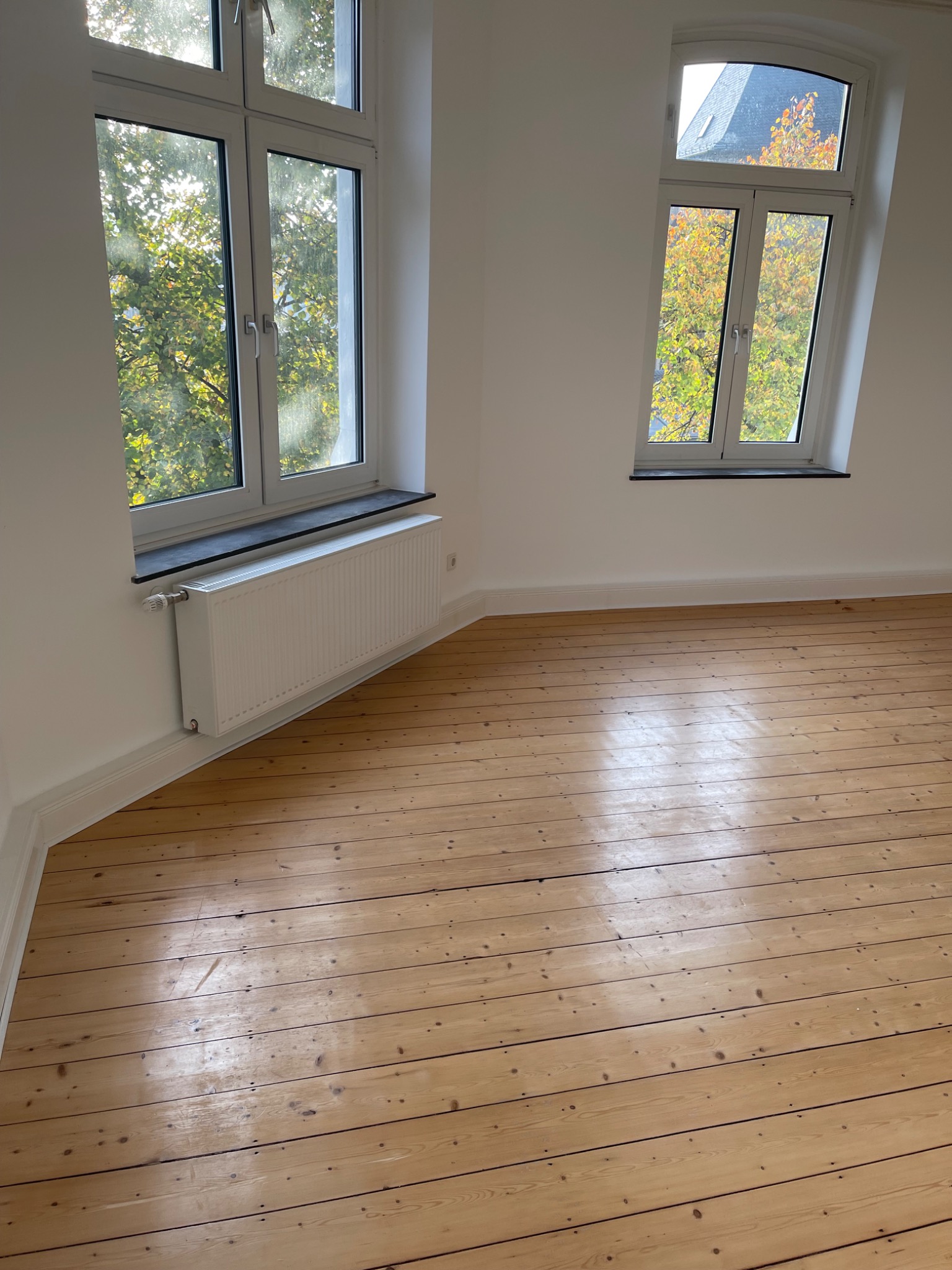 Pusty pokój z odnowioną drewnianą podłogą, białymi ścianami, dwoma oknami z widokiem na drzewa i grzejnikiem pod jednym z nich.
