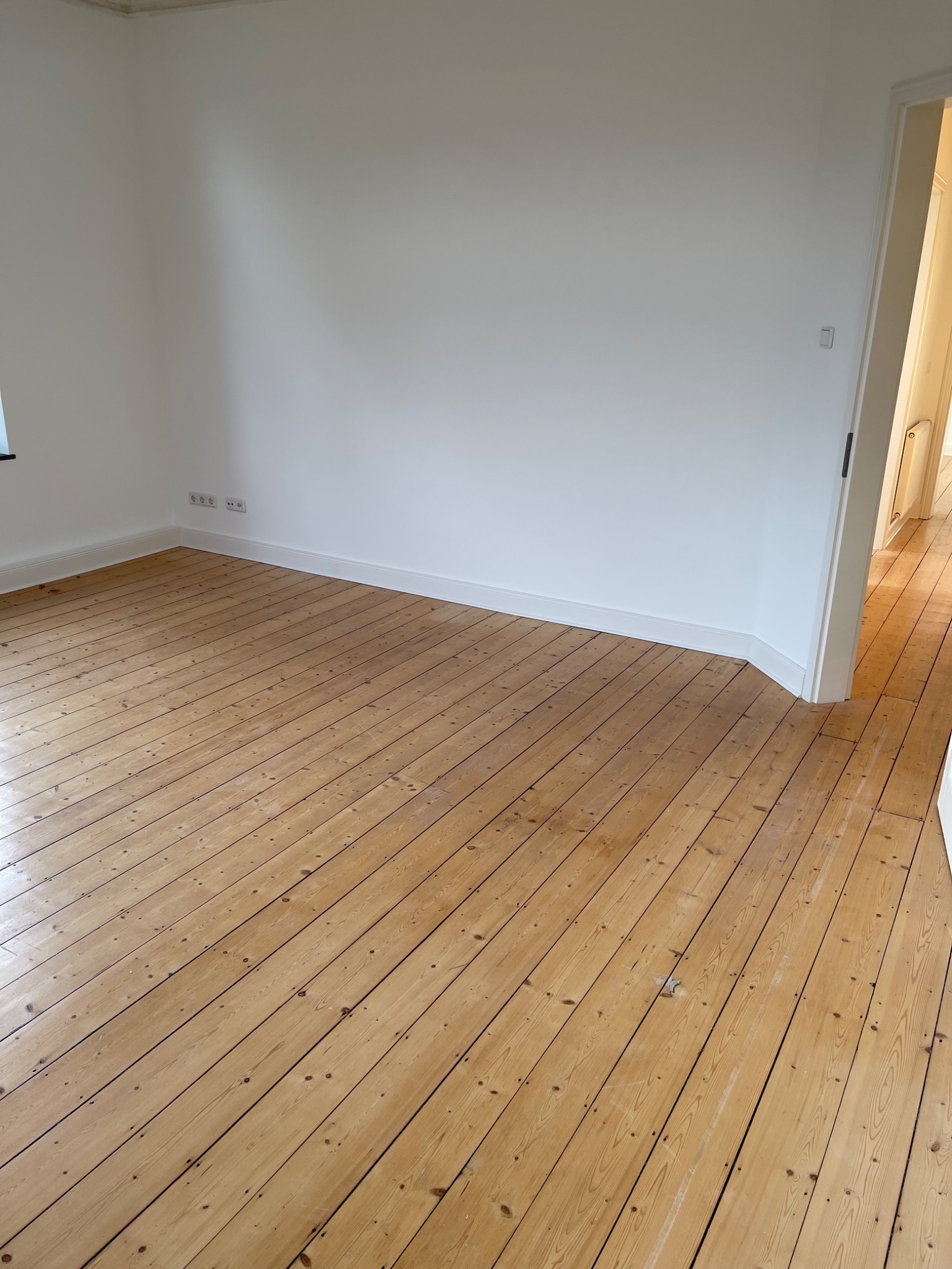 Pusta, jasna przestrzeń z drewnianą podłogą ułożoną ukośnie, białe ściany z listwami przypodłogowymi i gniazdkami elektrycznymi, widok na sąsiednie pomieszczenie przez otwarte drzwi.
