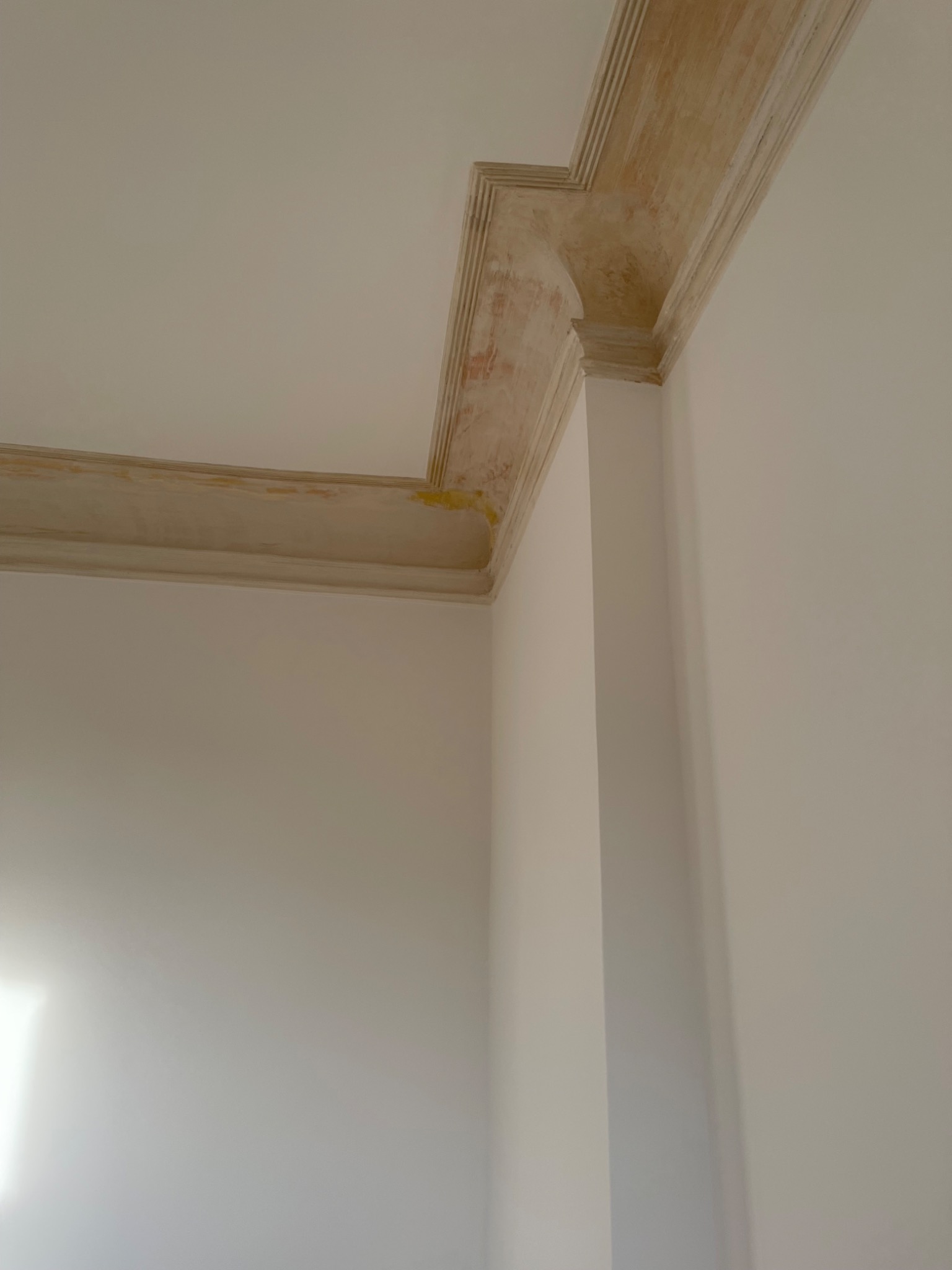 Narożnik pokoju z pomalowaną na beżowo sztukaterią sufitową z żółtymi przebarwieniami, na tle białych ścian po remoncie.