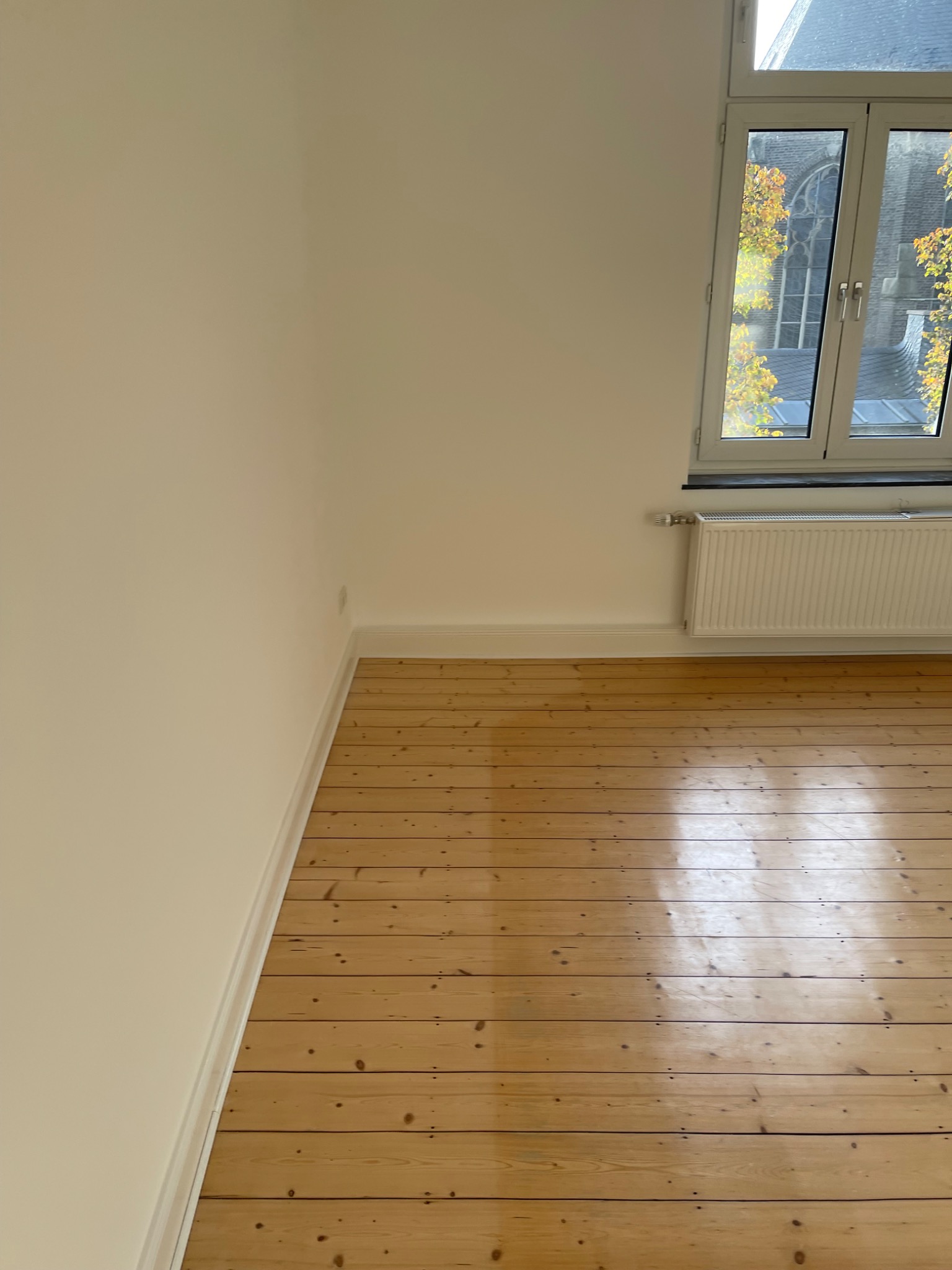 Jasne, odnowione wnętrze z drewnianą podłogą, świeżo pomalowanymi ścianami na kremowo i białym grzejnikiem pod oknem z widokiem na kościół w oddali.