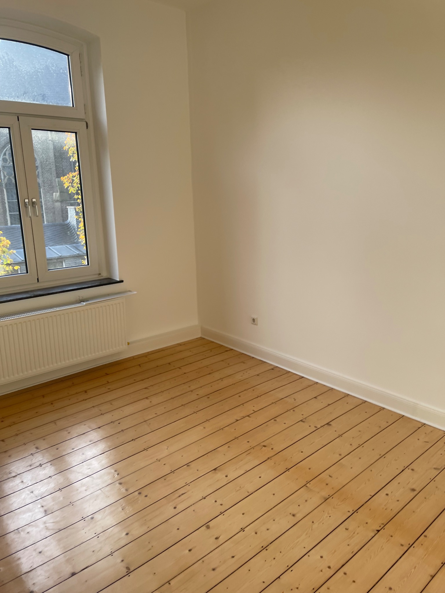 Pusty pokój z odnowioną drewnianą podłogą, świeżo pomalowanymi ścianami w kolorze ecru, białym grzejnikiem pod oknem z widokiem na jesienne drzewa i budynek z czerwonej cegły.