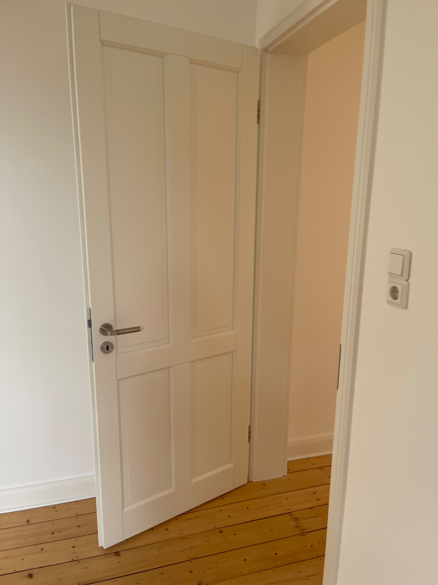 Otwarta, biała, płycinowa drzwi wewnętrzne z srebrną klamką, widoczne drewniane panele podłogowe i białe ściany, z gniazdkiem elektrycznym po prawej stronie.