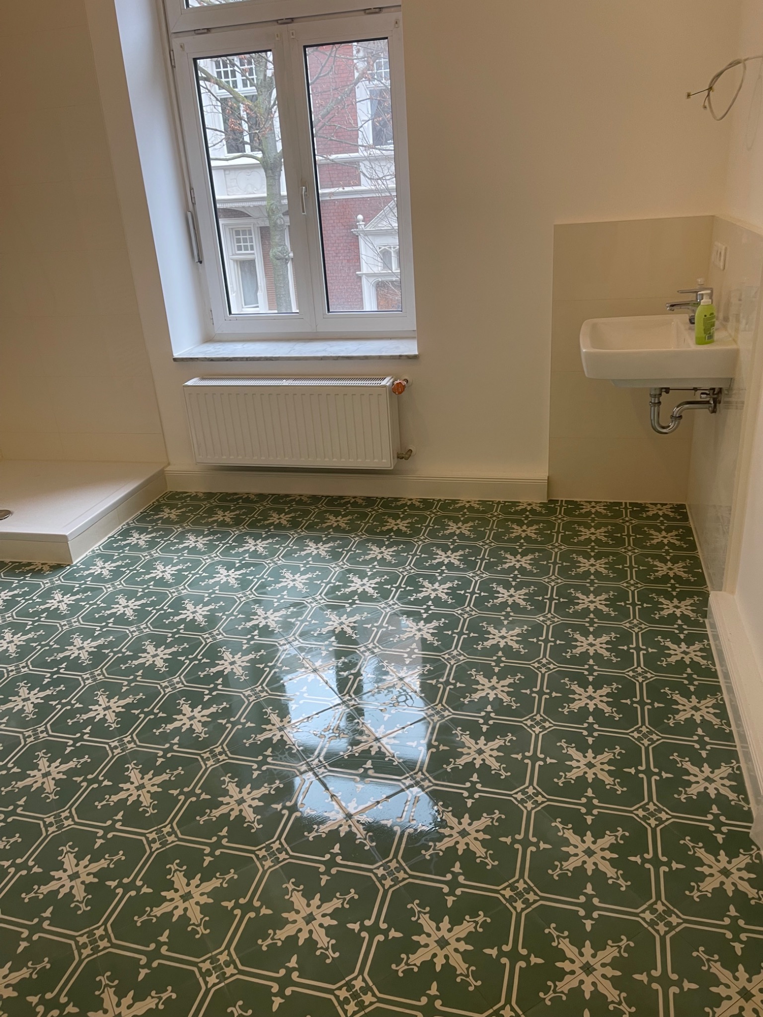 Łazienka po remoncie z nową, błyszczącą, zieloną podłogą w stylu retro z białym wzorem, umywalką i grzejnikiem pod oknem, częściowo widoczny brodzik.