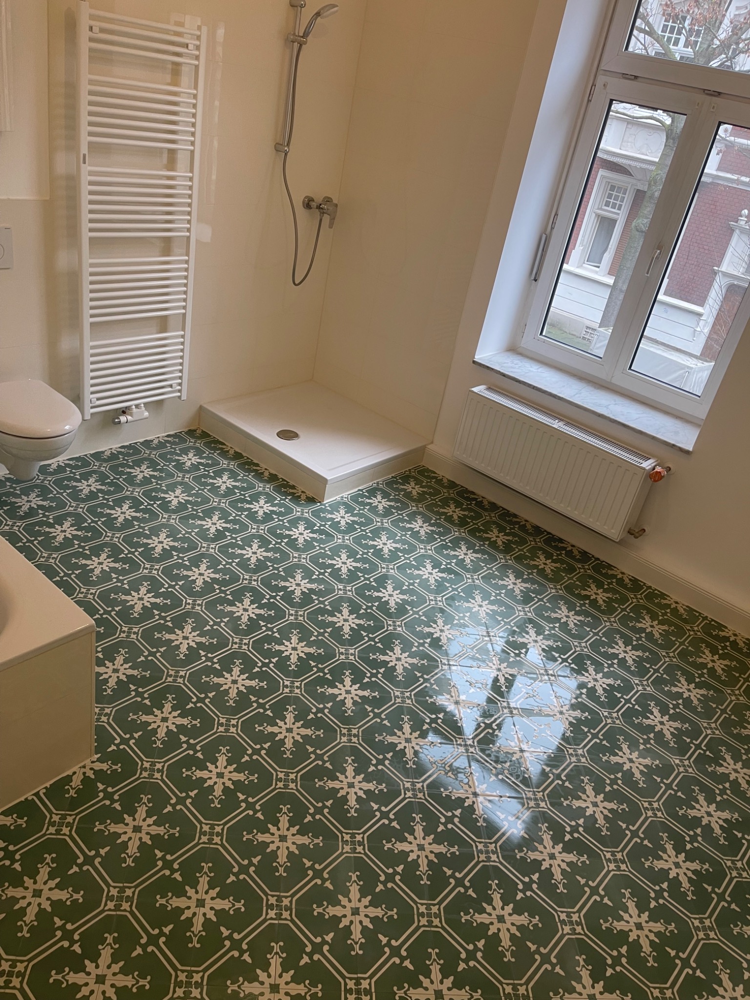 Wykończona łazienka z zieloną, wzorzystą podłogą, białą kabiną prysznicową, grzejnikiem drabinkowym i oknem z widokiem na zabudowę miejską.