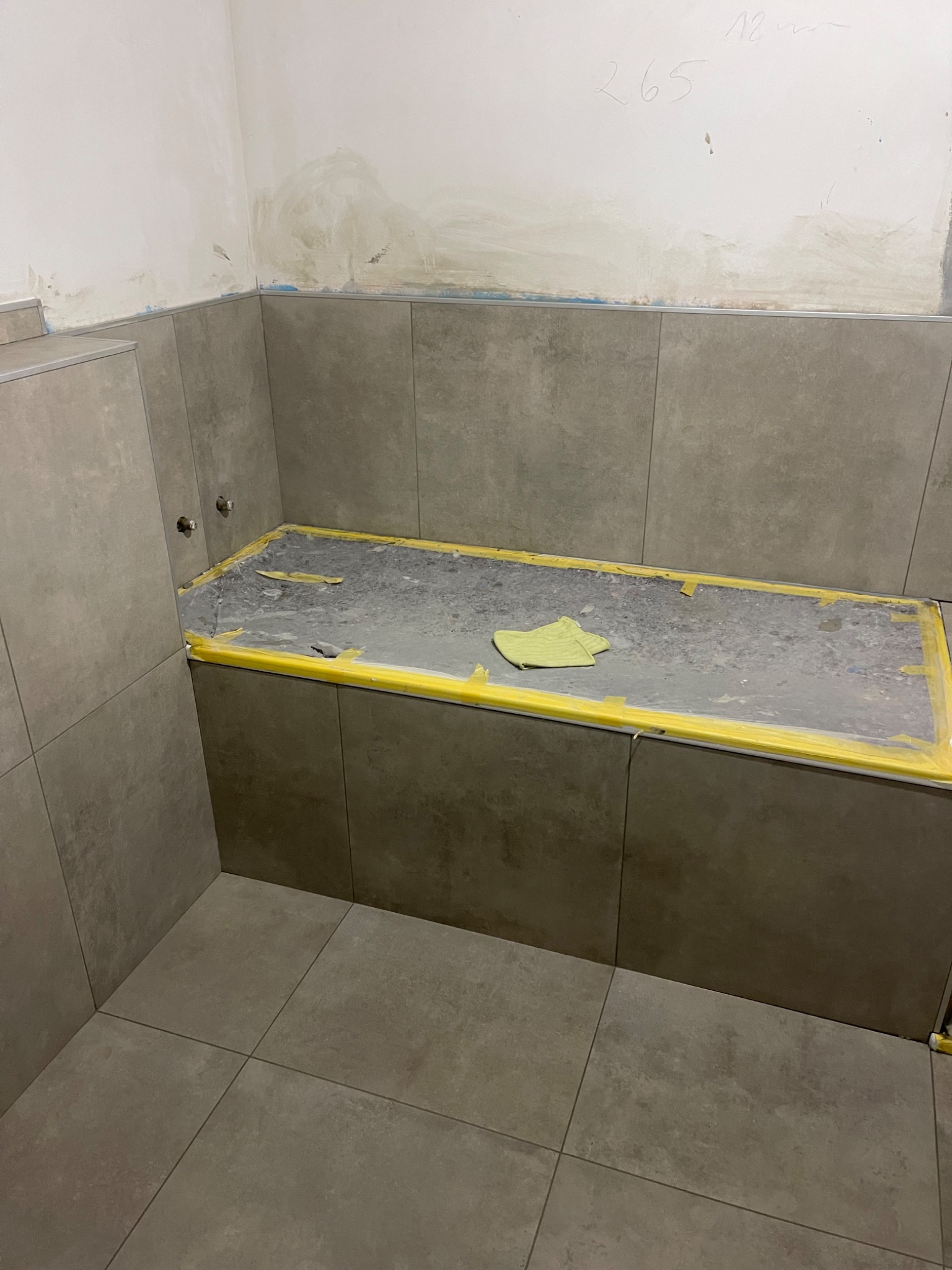 Wnętrze łazienki w trakcie remontu, widoczne szare płytki na ścianach i podłodze, zabezpieczona żółtą taśmą wanna, ślady po zaprawie na ścianie i odręczny napis '265' nad wanną.