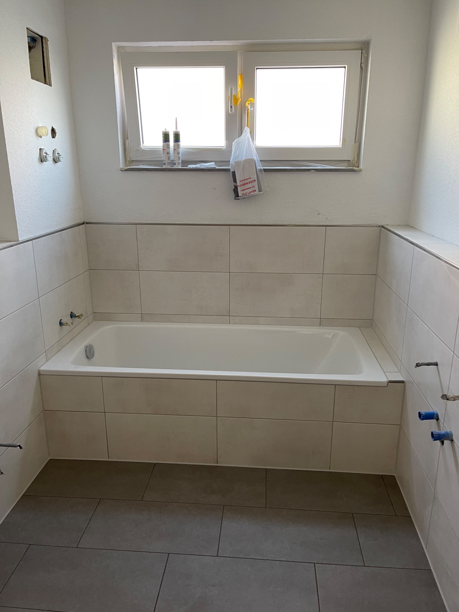 Wanna obudowana jasnymi płytkami w trakcie remontu łazienki. Widoczne wystające rury instalacji wodnej, szare płytki podłogowe, okno z tubami silikonu na parapecie i workiem z materiałami montażowymi.