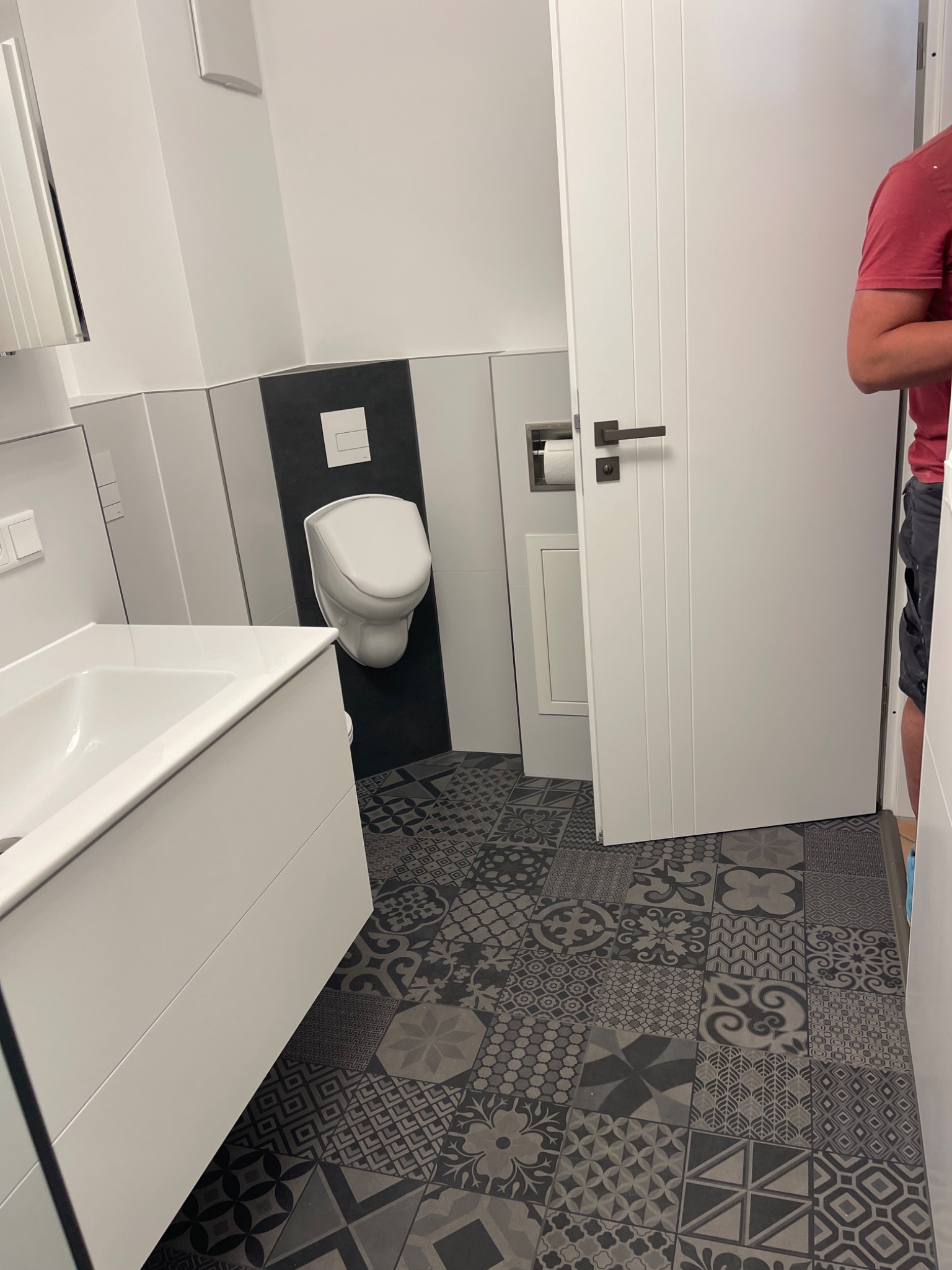 Wykończona łazienka z umywalką, wiszącym WC na czarnym tle i wzorzystymi, ciemnymi płytkami podłogowymi; widoczny fragment białych drzwi i osoby w czerwonej koszulce.