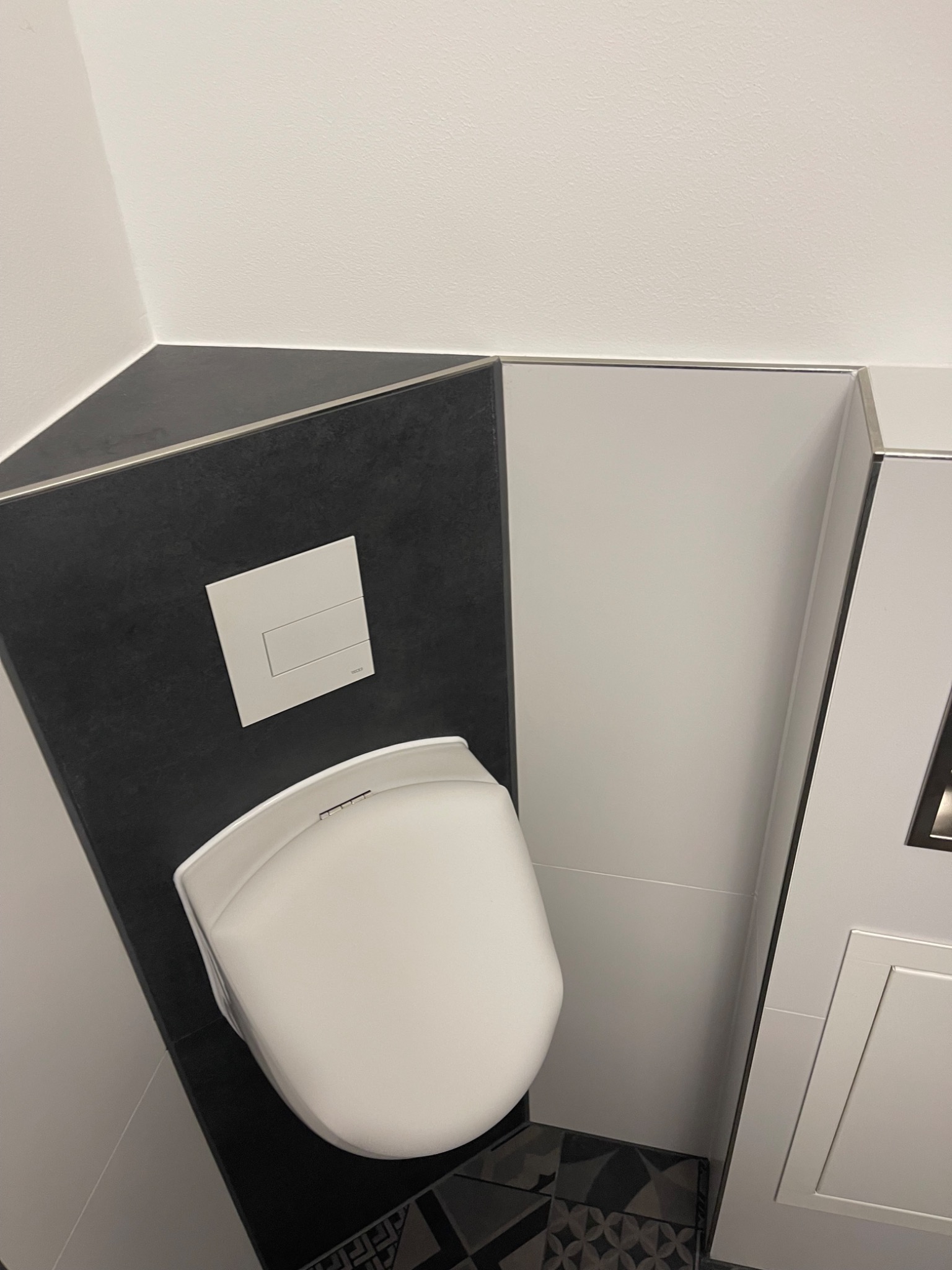 Nowoczesna łazienka z wiszącą toaletą, czarno-białe płytki na ścianach i podłodze, minimalistyczny design, przycisk spłukiwania Geberit na czarnej ścianie.