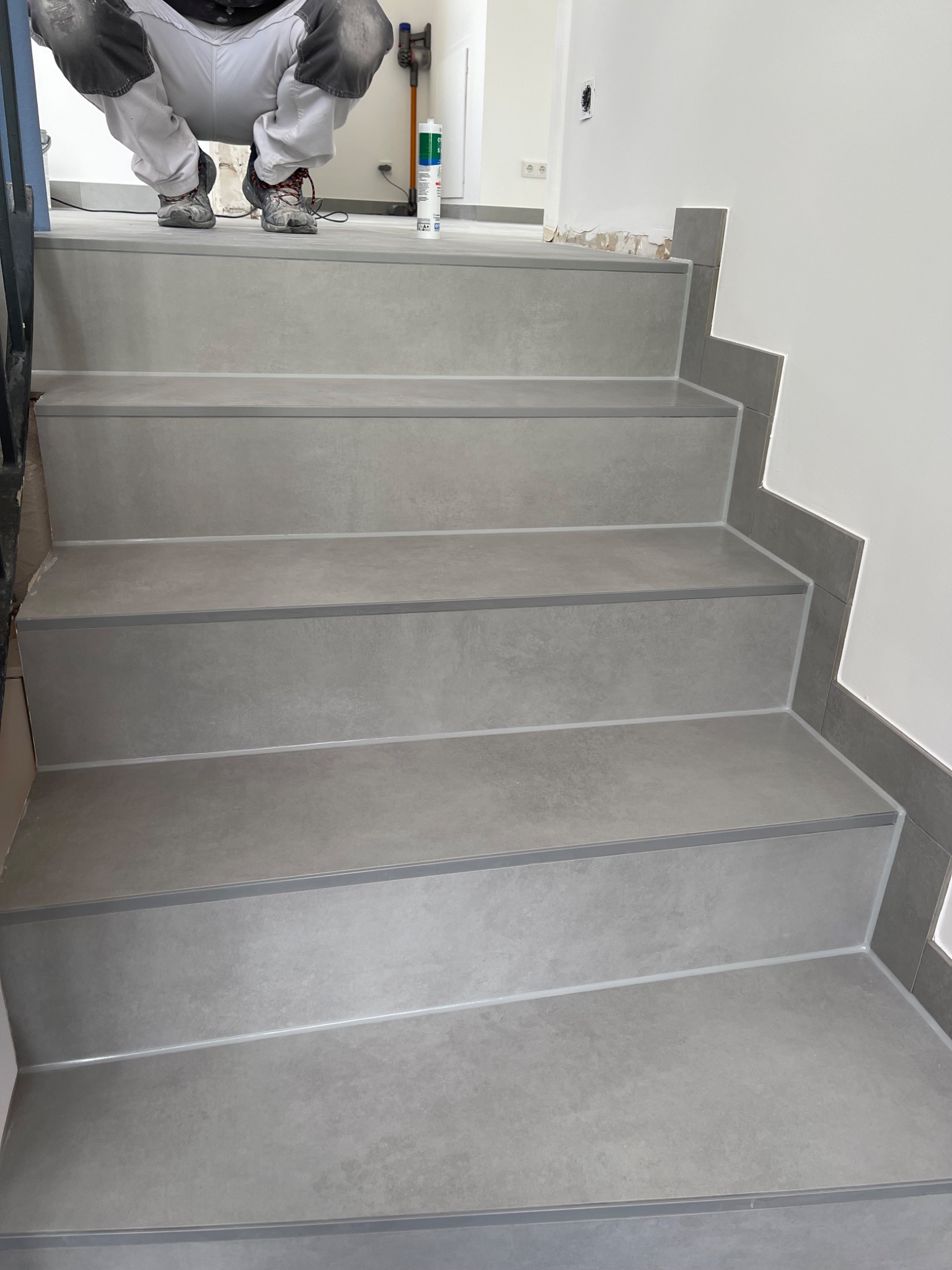Wykończone schody z szarych płytek, widoczny silikon na krawędziach, pracownik w ubrudzonych spodniach i butach, tuba silikonu i odkurzacz w tle.