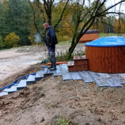 Montaż schodów z płyt kamiennych prowadzących do drewnianej balii w Łukcie, widoczny pracownik w odzieży roboczej i częściowo ułożony taras z płyt kamiennych.