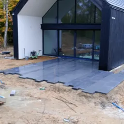 Układanie ciemnoszarych płyt tarasowych przy nowoczesnym domu z dużymi przeszkleniami; widoczne narzędzia i częściowo ułożona powierzchnia tarasu na gruncie.