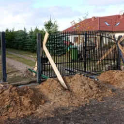 Montaż czarnej, metalowej bramy wjazdowej na posesję, z drewnianymi podporami tymczasowymi i usypanym gruntem wokół słupków, widok na dom z czerwoną dachówką i zadbany ogród.