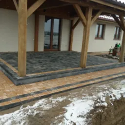 Zadaszony taras z kostki brukowej w odcieniach szarości i beżu, z obrzeżami z ciemnej kostki. Widoczne drewniane słupy podtrzymujące zadaszenie, fragment ośnieżonego terenu i elewacji budynku w tle.