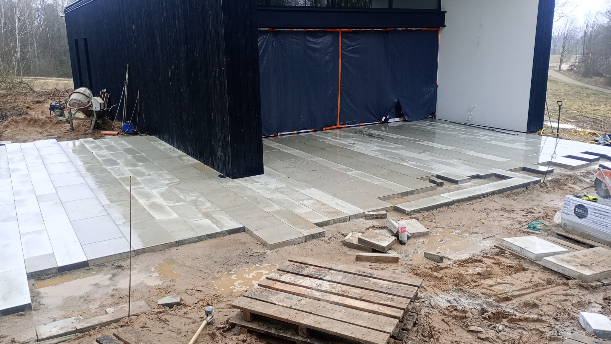 W trakcie układania szarej kostki brukowej na posesji z nowoczesnym budynkiem o czarno-białej elewacji; widoczny mikser do betonu, paleta z kostką oraz narzędzia budowlane.