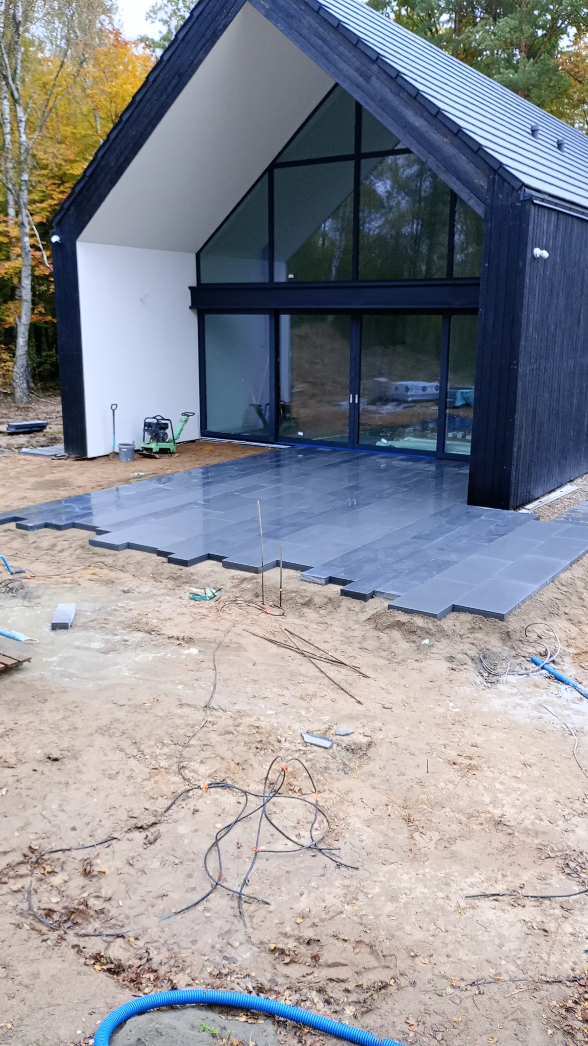 Układanie ciemnoszarej kostki granitowej tarasowej przy nowoczesnym domu z dużymi przeszkleniami, widoczna ubijarka w tle.