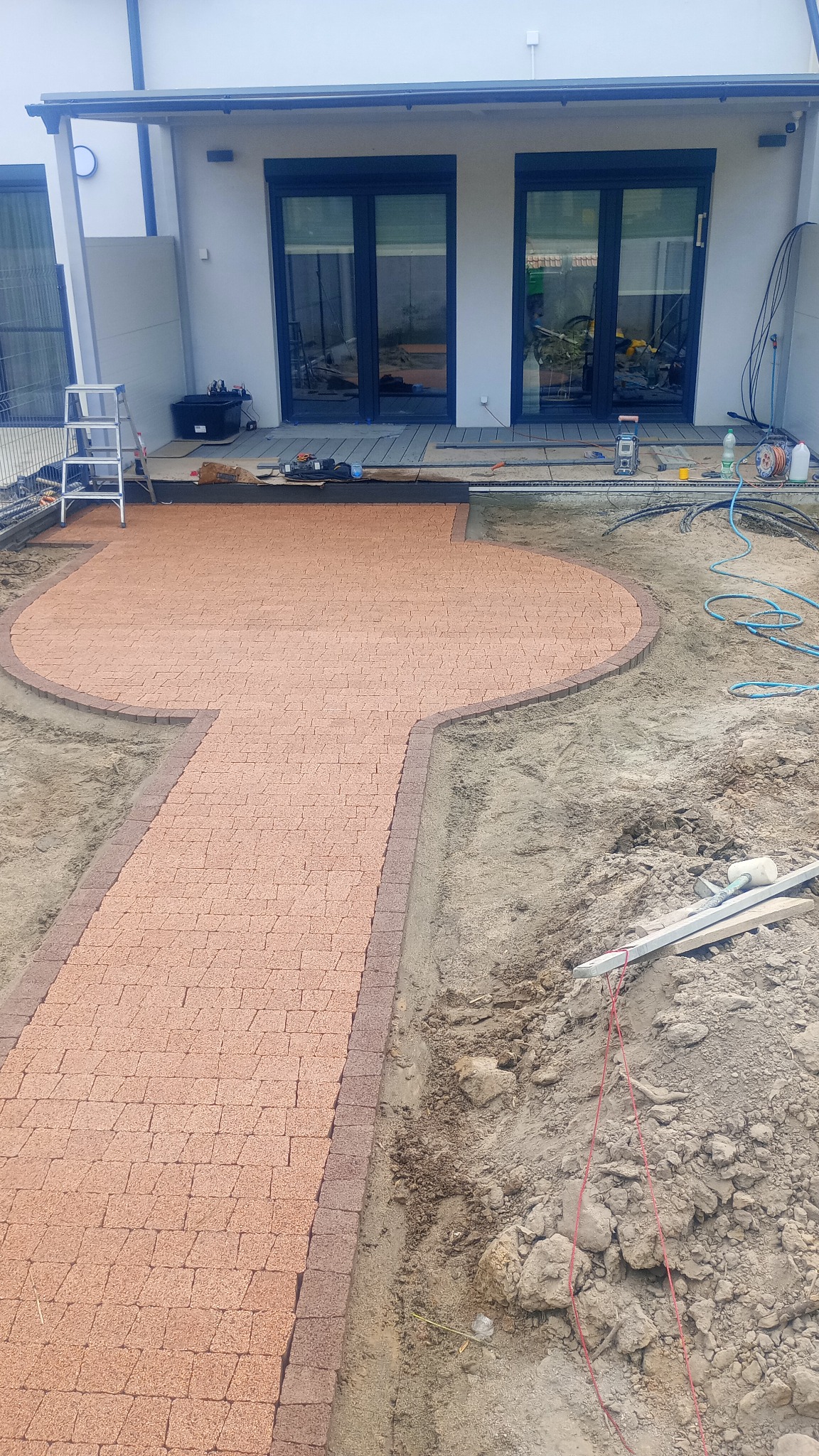 Czerwono-brązowa kostka brukowa ułożona w kształcie ścieżki i okrągłego tarasu przed domem, widoczne narzędzia i materiały budowlane, teren w trakcie prac ziemnych.