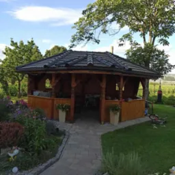 Drewniana altana z ciemną dachówką, otoczona kwiatami i zielenią, z widocznym stołem i krzesłami wewnątrz, ścieżka z kostki prowadzi do wejścia.