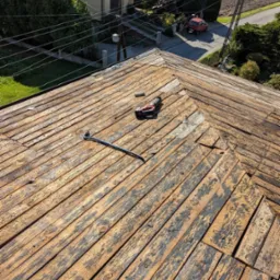 Demontaż starego poszycia z desek na dachu domu jednorodzinnego, widoczne narzędzia: wkrętarka akumulatorowa i łom.