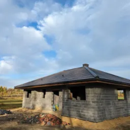 Surowy budynek jednorodzinny z ciemną dachówką ceramiczną, widoczne bloczki betonowe ścian i otwory okienne, na ziemi porozrzucane fragmenty dachówek, tło stanowi błękitne niebo z chmurami...