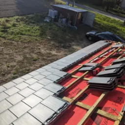 Montaż szarej dachówki na czerwonej membranie, widoczne drewniane łaty, w tle budynek gospodarczy, samochód i zieleń.