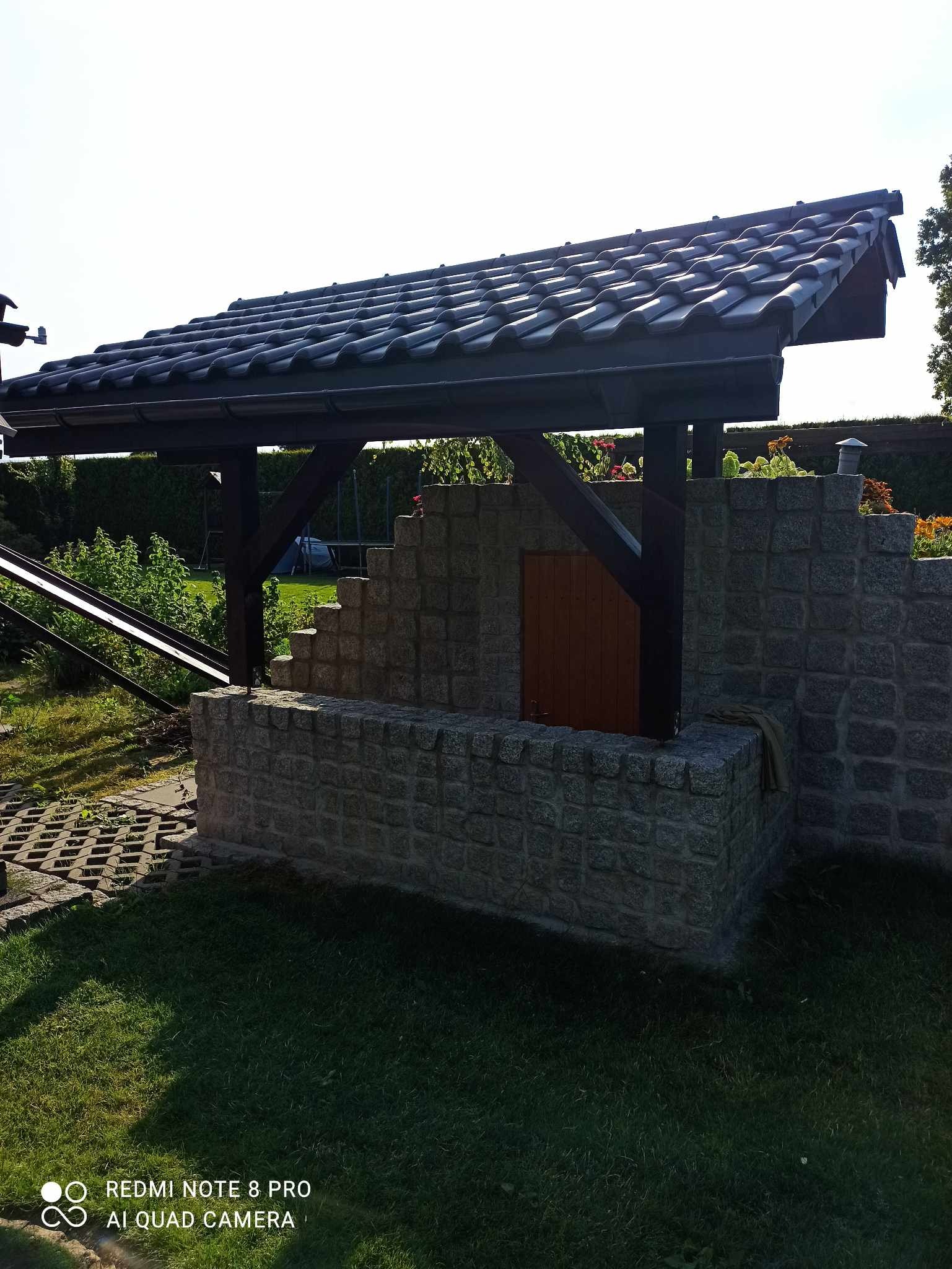 Murowana konstrukcja z kamienia, z drewnianym zadaszeniem pokrytym ciemną dachówką, umieszczona w ogrodzie.