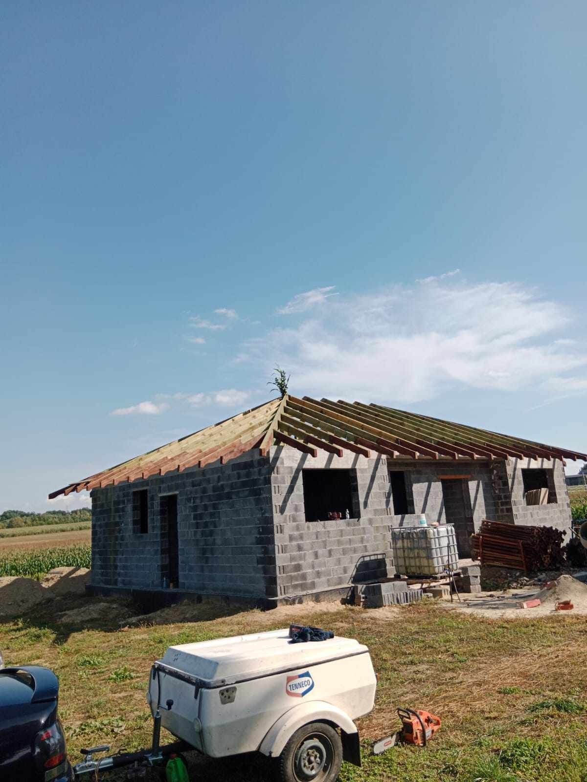 Surowy budynek w trakcie budowy z częściowo ułożoną konstrukcją drewnianą dachu, widoczny także wózek transportowy i piła łańcuchowa.