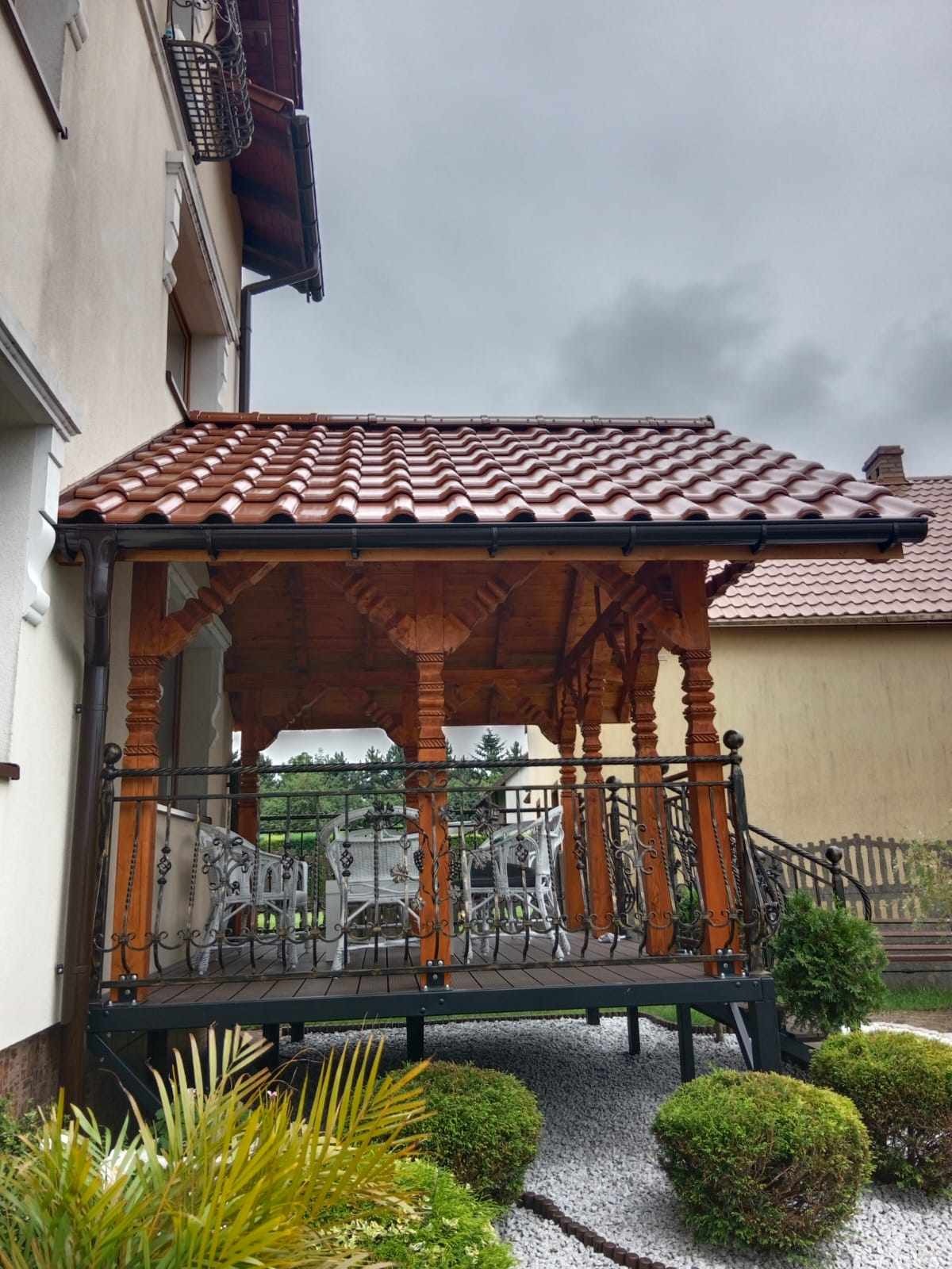 Drewniana altana z brązowym dachem z dachówki ceramicznej, ozdobnymi słupami i czarną, metalową balustradą, otoczona zielenią i białym żwirem.