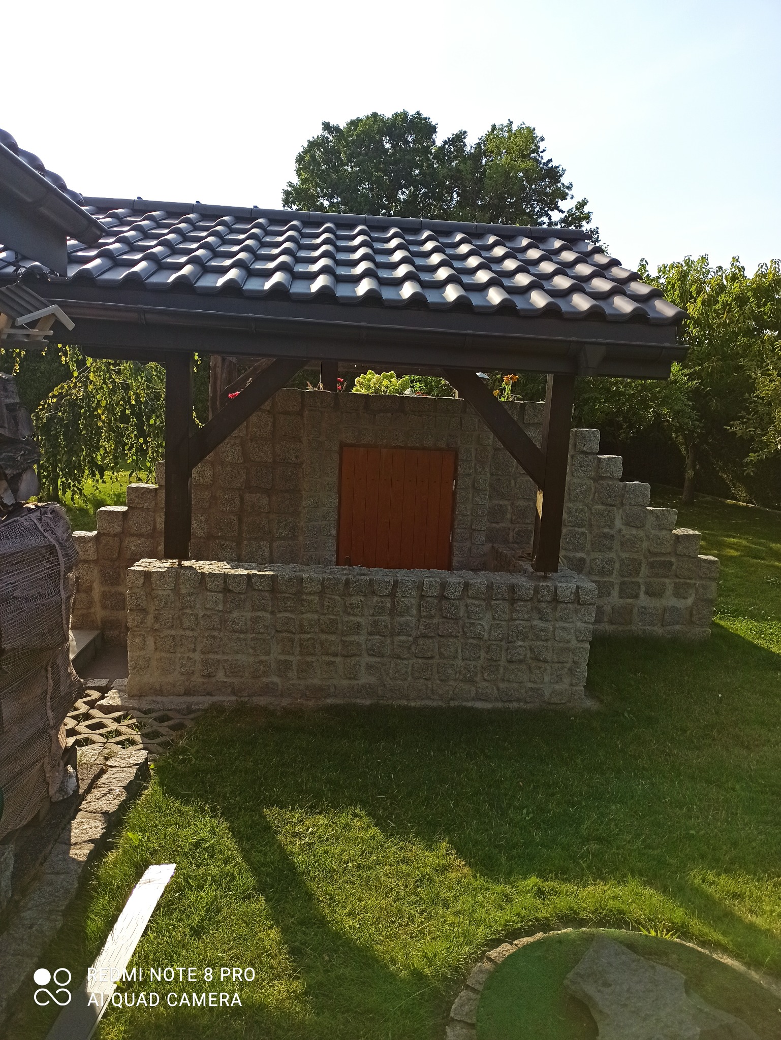 Kamienna altana z drewnianą konstrukcją dachu krytą ciemną dachówką ceramiczną, otoczona zielonym trawnikiem i drzewami w słoneczny dzień.
