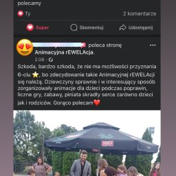 Animacyjna rEWELAcja - Animator w szarej marynarce nadzoruje grupę dzieci bawiących się na trawie podczas imprezy plenerowej, widoczny parasol reklamowy.