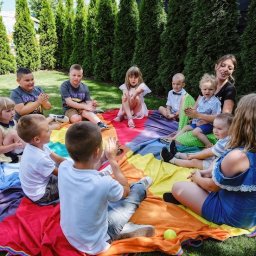 Animacyjna rEWELAcja - Grupa dzieci siedzi na kolorowej chuście na trawie podczas zajęć z animatorką, w tle rząd wysokich tuj.