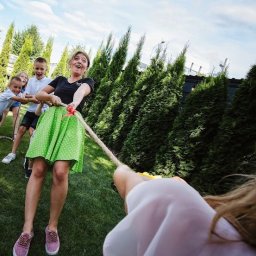 Animacyjna rEWELAcja - Grupa dzieci i dorosła kobieta w zielonej spódnicy grają w przeciąganie liny na trawniku, tło stanowią wysokie tuje.