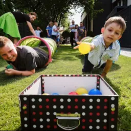 Chłopcy czołgają się przez tunele na trawie, celując piłkami do pudełka w kropki podczas zabawy z animatorem w ogrodzie.