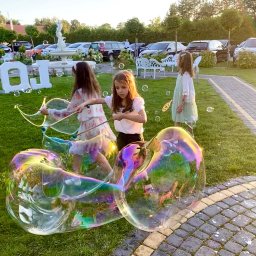 Animacyjna rEWELAcja - Trzy dziewczynki w Sochaczewie bawią się na trawniku, tworząc ogromne bańki mydlane za pomocą specjalnych kijków i sznurków. W tle widać fontannę, białe dekoracje i zaparkowane samochody.