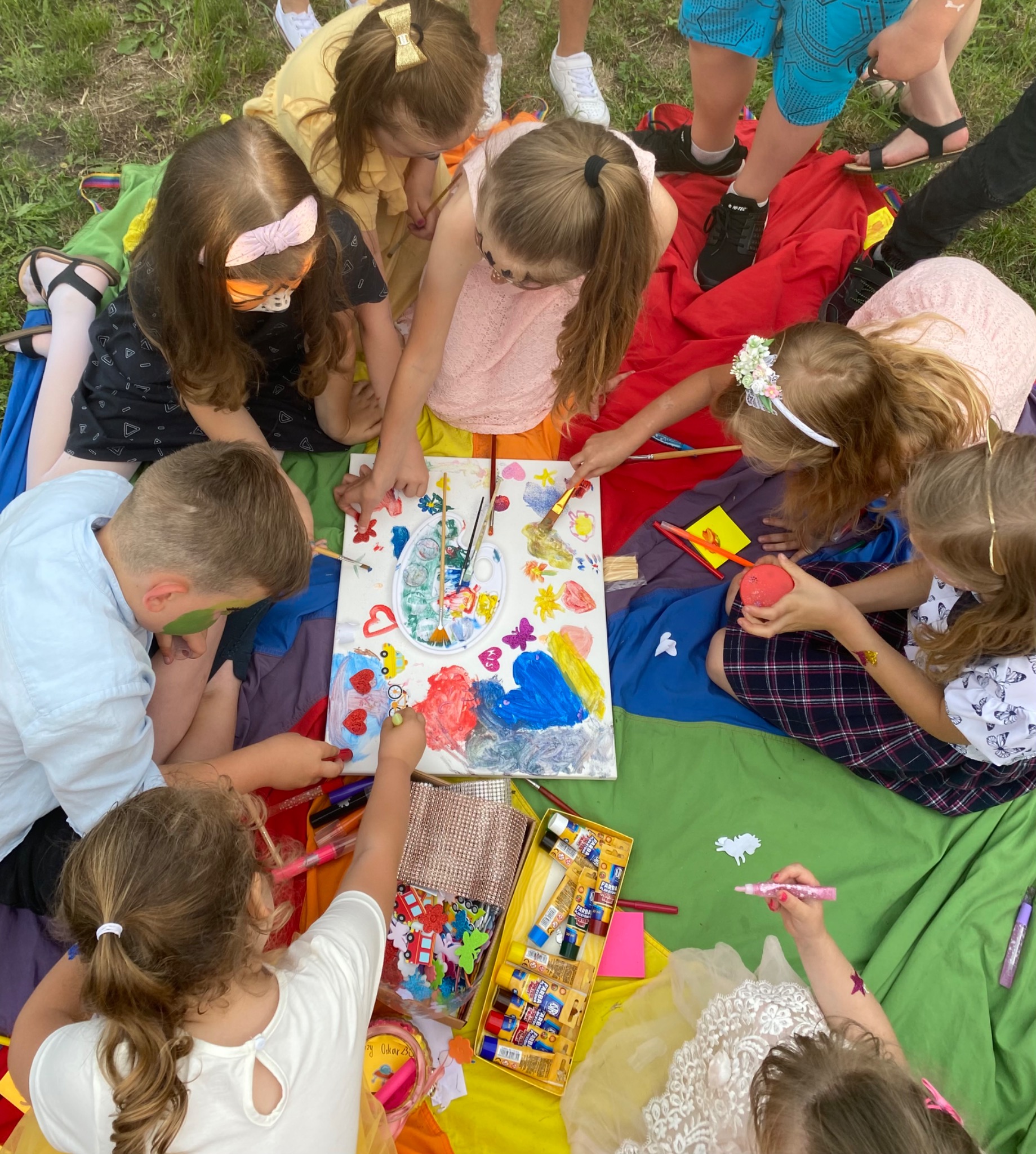 Grupa dzieci siedzących na kolorowym kocu, wspólnie malujących farbami na płótnie, z widocznymi tubkami farb, pędzlami i paletą.