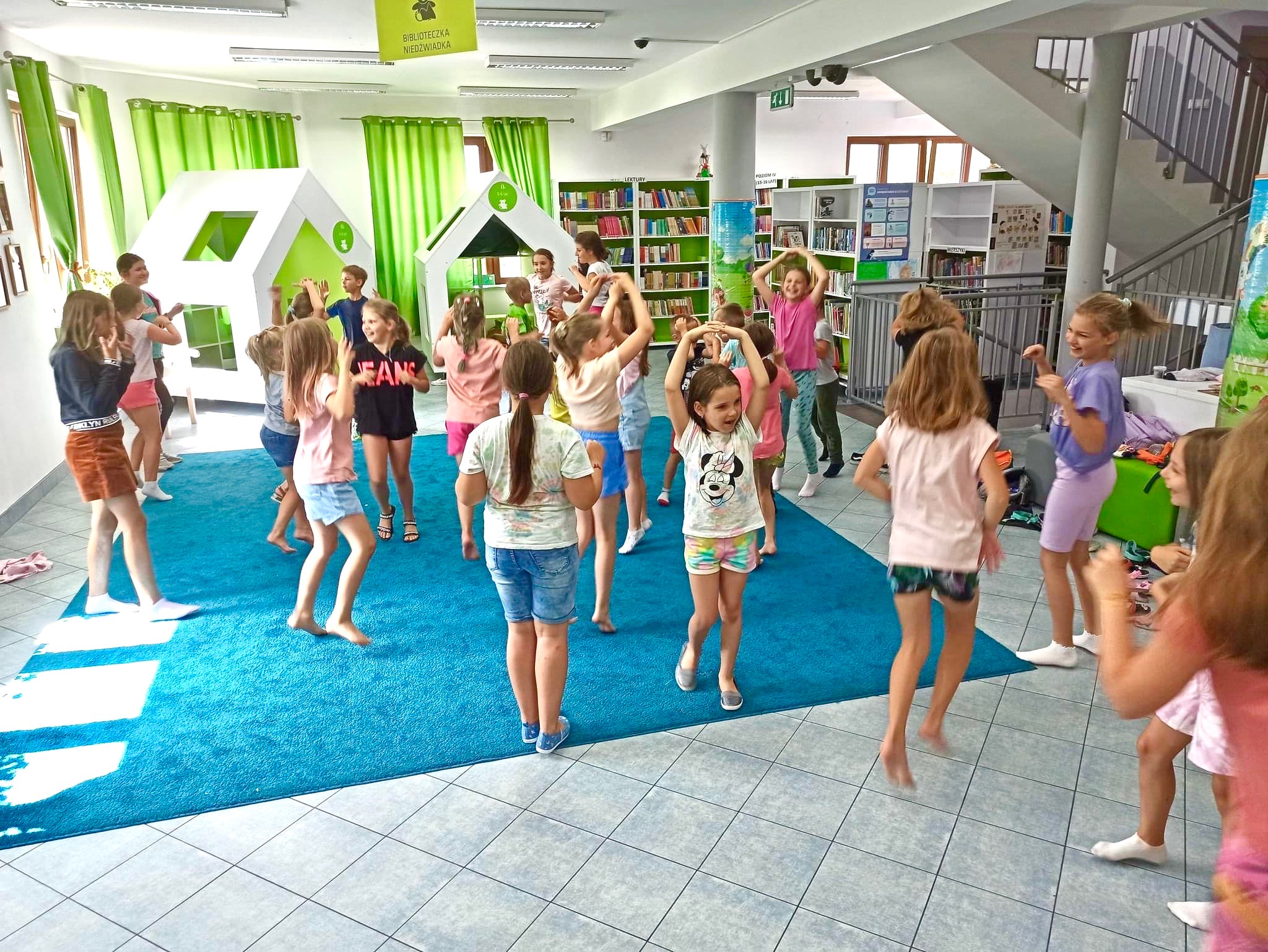 Grupa dzieci w bibliotece tańcząca na niebieskim dywanie, z białymi domkami zabaw w tle i regałami z książkami.