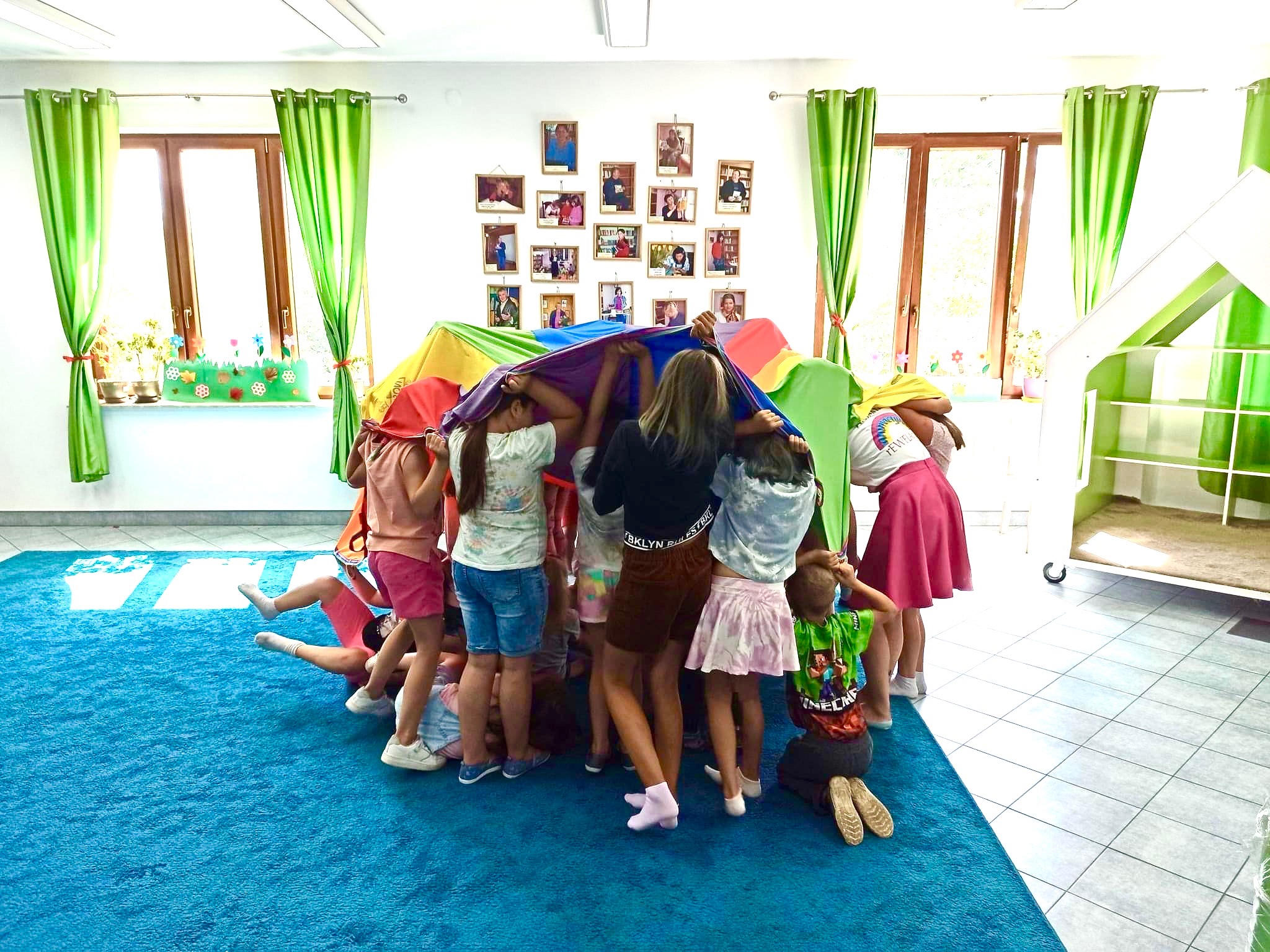 Grupa dzieci bawi się pod kolorową chustą w jasnym pomieszczeniu z zielonymi zasłonami i zdjęciami na ścianie.
